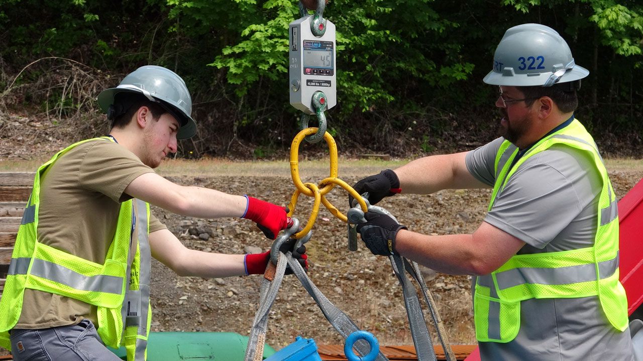 Two trainees wearing hard hats and safety vests collaborate to measure weight tension using a digital dynamometer as part of an ITI rigging course.